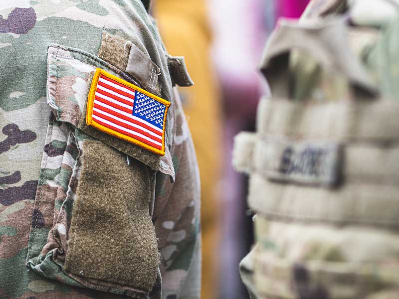 Close up of two military uniform sleeves: the one in the back is blurred out, and the one in front is not blurred out and has a patch of the American Flag